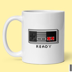 Puede incluir: Taza de cerámica blanca con una imagen pixelada en negro y gris de un mando de videojuego clásico y la palabra "READY" en texto pixelado.
