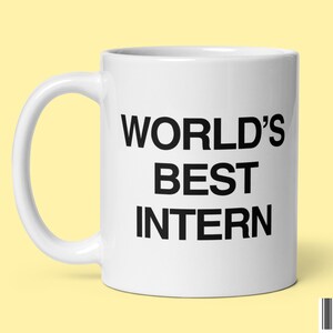 May include: White ceramic mug with the text "WORLD'S BEST INTERN" in black, with a curved handle. The mug is set against a light yellow background.
