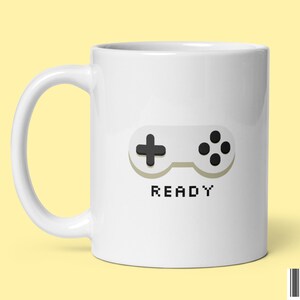 Puede incluir: Taza de cerámica blanca con una ilustración en blanco y negro de un mando de videojuego y la palabra "READY" en texto pixelado.