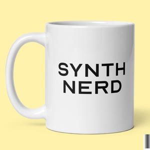 Może przedstawiać: Biały kubek ceramiczny z czarnym napisem "SYNTH NERD".