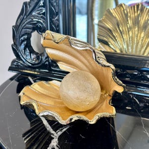 May include: A decorative seashell-shaped dish with a gold finish, holding a light-colored sphere. The dish has a silver trim and sits on a black surface. A black, ornate frame is in the background.