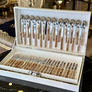 Puede incluir: Un juego completo de cubiertos en una caja blanca. El juego incluye cucharas, cuchillos y tenedores. Los mangos tienen un diseño similar al bambú con detalles dorados. Los cubiertos están ordenados cuidadosamente dentro de la caja.