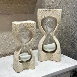 Op de afbeelding: Twee decoratieve zandlopers met een getextureerde, gebroken witte steenachtige buitenkant. De zandlopers hebben een helder glazen midden gevuld met wit zand. De zandlopers zijn verschillend van grootte en staan op een wit oppervlak.
