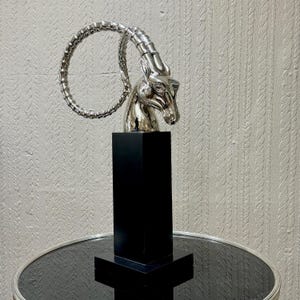 May include: A silver-colored sculpture of an animal head with curved horns, mounted on a black rectangular base. The sculpture is placed on a round, black glass table. The background is a textured, light-colored wall.