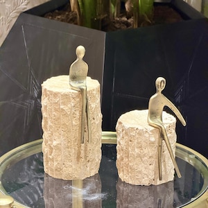 Könnte beinhalten: Zwei dekorative Skulpturen. Jede zeigt eine kleine, goldfarbene Figur, die auf einem strukturierten, beigefarbenen Steinzylinder sitzt. Die Skulpturen werden auf einer reflektierenden Oberfläche mit dunklem, geometrischem Hintergrund präsentiert.