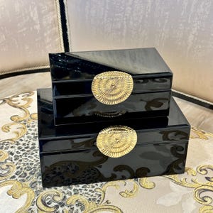 Pode incluir: Duas caixas decorativas pretas e brilhantes empilhadas uma sobre a outra. Cada caixa tem uma alça circular dourada. As caixas estão sobre uma superfície estampada.