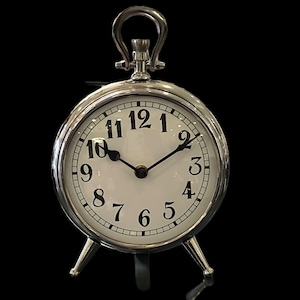 May include: A silver-colored, round analog clock with a white face and black numerals. The clock has a handle at the top and three small legs. The hands point to approximately 10:09.