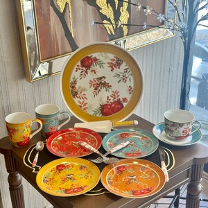 May include: A collection of decorative ceramic tableware featuring a fall harvest theme. Includes plates, mugs, a teacup and saucer, and serving utensils. The plates have a bird and pomegranate design with red, yellow, and orange accents.