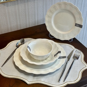 Op de afbeelding: Een crèmekleurige servies set met een geschulpte rand en een kralenrand. De set bevat een dinerbord, een saladebord, een kom, een lepel, een mes en een vork. De borden en de kom zijn gestapeld op een placemat met een gouden rand.