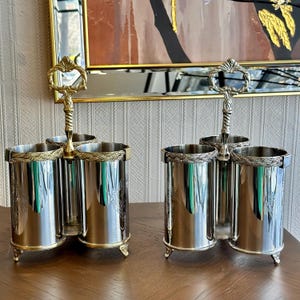 Puede incluir: Dos especieros plateados con asas y adornos dorados. Cada uno tiene tres compartimentos cilíndricos. Las asas son ornamentadas y las bases tienen pequeños pies decorativos. Los especieros están sobre una superficie de madera.