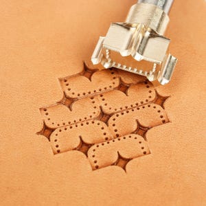 May include: A silver-colored metal leather stamping tool positioned above a tan leather surface. The tool features a multi-pointed star design that has been pressed into the leather, creating a repeating pattern of indented star shapes.