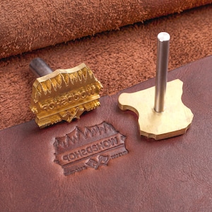 Op de afbeelding: Twee messing lederstempels met metalen handvatten. Eén stempel heeft een reliëfontwerp van een bos, twee gekruiste bijlen en de tekst WOODSHOP. De andere is een platte messing plaat met een verticaal metalen staafhandvat.