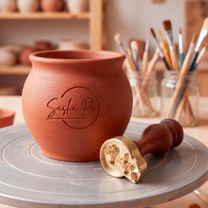 Puede incluir: Una olla de cerámica de color terracota con el texto "Sasha Pi" en un diseño circular. Un sello de latón con mango de madera y diseño floral está al lado de la olla. Pinceles en el fondo.