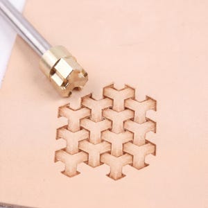 May include: A leathercraft tool with a geometric pattern stamp. The stamp is made of brass and has a hexagonal shape. The pattern is stamped onto a piece of light brown leather, creating a repeating design.