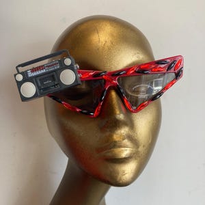May include: Red and black patterned sunglasses with a small, black boombox attached. The sunglasses have a unique, angular shape and are displayed on a gold mannequin head. The boombox is a miniature replica.