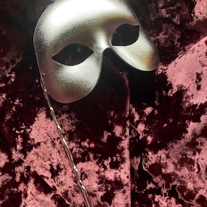 May include: A silver masquerade mask with a long chain attached. The mask is lying on a burgundy velvet fabric.