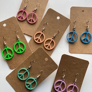 Op de afbeelding: Een verzameling kleurrijke peace-teken oorbellen in roze, groen, oranje, blauw en paars. Elke paar wordt gepresenteerd op een bruine kaart met een zilveren haak. De oorbellen zijn een vrolijk accessoire.