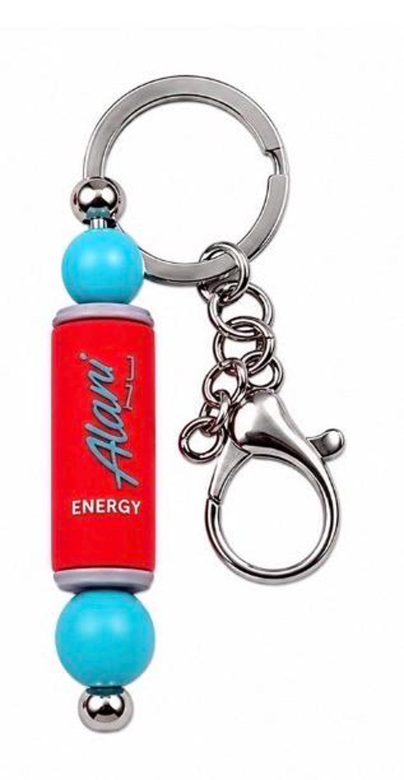 Puede incluir: Un llavero de metal con un dije cil&iacute;ndrico rojo que tiene el texto Alani ENERGY impreso en blanco y gris. El dije est&aacute; acentuado por dos cuentas esf&eacute;ricas de color turquesa brillante y dos cuentas de metal plateado m&aacute;s peque&ntilde;as en una argolla.