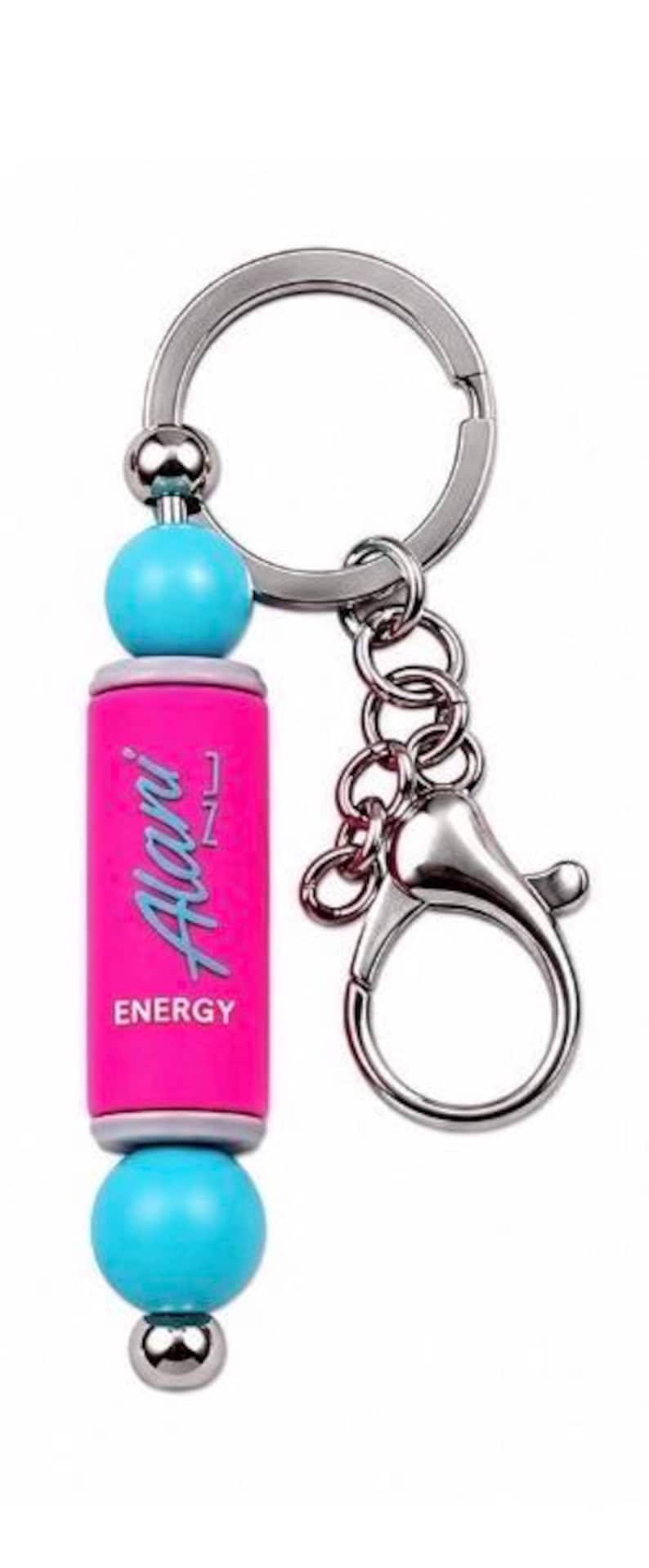 Puede incluir: Un llavero con un dije cil&iacute;ndrico de color rosa brillante con el texto Alani Energy impreso en una fuente cursiva azul claro. El dije est&aacute; acentuado por dos cuentas redondas de color turquesa y una peque&ntilde;a cuenta plateada, unido a un anillo met&aacute;lico.