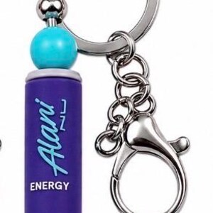 Puede incluir: Un llavero con un dije cil&iacute;ndrico morado con el texto Alani ENERGY impreso en blanco y azul. El dije est&aacute; acentuado por dos cuentas esf&eacute;ricas de color turquesa brillante y una peque&ntilde;a cuenta plateada en la base, unido a una argolla de metal plateado.