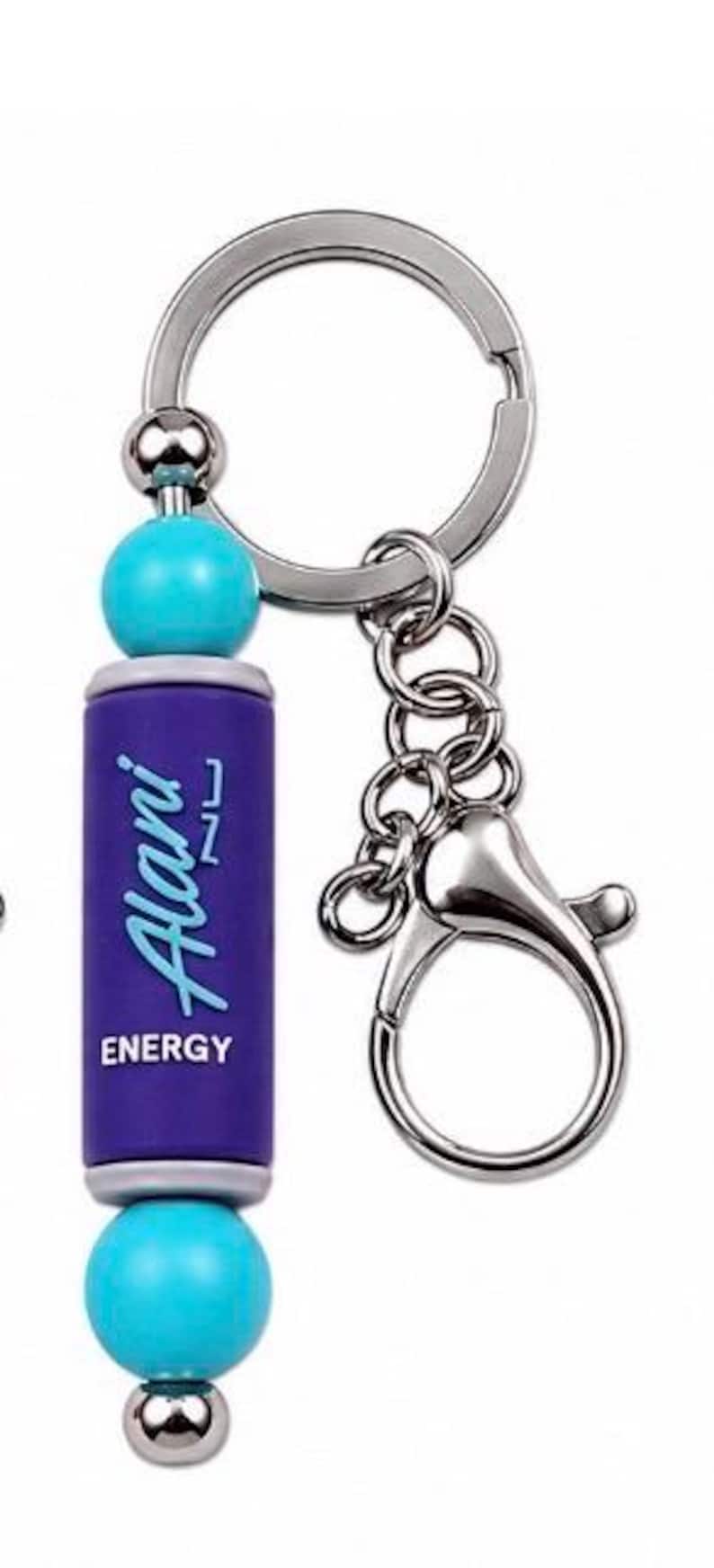 Puede incluir: Un llavero con un dije cil&iacute;ndrico morado con el texto Alani ENERGY impreso en blanco y azul. El dije est&aacute; acentuado por dos cuentas esf&eacute;ricas de color turquesa brillante y una peque&ntilde;a cuenta plateada en la base, unido a una argolla de metal plateado.