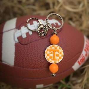 May include: A silver keychain with orange beads and a circular charm featuring a white and orange checkerboard design with a large orange letter "T". The keychain is displayed in front of a brown football.