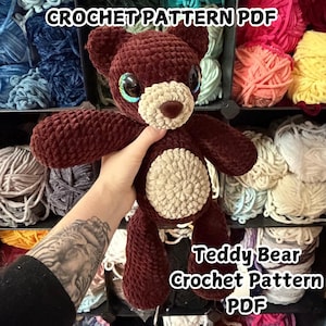 May include: A handmade, dark brown teddy bear with a cream-colored muzzle and belly, and large, expressive eyes. The bear is crocheted and held in front of a shelf filled with yarn. Text on the image reads "CROCHET PATTERN PDF" and "Teddy Bear Crochet Pattern PDF."