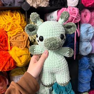 May include: A pale green crocheted giraffe with dark green detailing on its ears, snout, and feet. The giraffe has large black eyes and is made of soft, textured yarn. The background features colourful skeins of yarn.