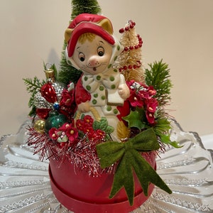 May include: A vintage Christmas decoration featuring a ceramic cat figurine wearing a red hat and scarf with green polka dots. The cat is surrounded by miniature Christmas trees, ornaments, and red flowers, all set in a red round box with a green bow.