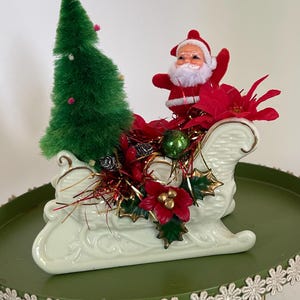 May include: A white ceramic sleigh filled with a green bottle brush tree, a small Santa Claus figurine, red poinsettia flowers, green holly leaves, and metallic tinsel. The sleigh features a textured basket-weave pattern and gold trim accents.