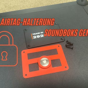 Könnte beinhalten: Ein roter Plastik-AirTag-Halter mit einem Ausschnitt für einen AirTag. Der Halter ist an einer schwarzen Oberfläche befestigt mit dem Text "AIRTAG-HALTERUNG SOUNDBOKS GEN.4" und einer schwarzen Platte mit dem Text "TEAM ID 292".