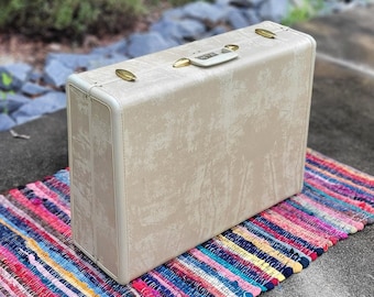 May include: A vintage off-white suitcase with a gold metal handle and a textured surface. The suitcase is sitting on a colorful striped rug.