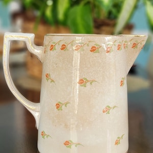 May include: A white ceramic pitcher with a gold rim and a floral design featuring orange flowers and green leaves. The pitcher has a handle and a spout.