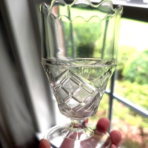 May include: A clear glass goblet featuring a scalloped rim and a textured bowl with a geometric diamond pattern. The stem connects to a circular base, and the glass has a slight green tint, showcasing a pressed glass design with clear transparency.