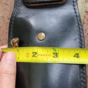May include: Black leather holster with a brass button and a brown leather strap. The holster is approximately 7.6 cm wide. A yellow tape measure is used to measure the width of the holster.