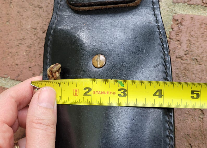May include: Black leather holster with a brass button and a brown leather strap. The holster is approximately 7.6 cm wide. A yellow tape measure is used to measure the width of the holster.