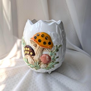 May include: White ceramic planter with a whimsical mushroom design. The planter features a large orange mushroom with black spots, a brown mushroom, and a pink mushroom, all surrounded by green leaves. The planter has a scalloped rim.