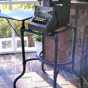 May include: A black vintage cash register on a black metal stand with three legs. The register has a paper roll and a display with the word "Carlton" on it. The stand is on a brick floor.