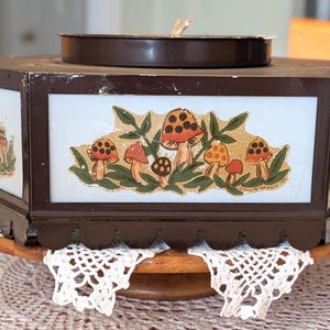 May include: A vintage, hexagonal, brown metal box with glass panels featuring mushroom illustrations. The box sits on a wooden stand with a scalloped edge and white lace doilies. A circular lid sits on top.