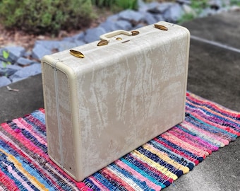 May include: A vintage white suitcase with gold hardware. The suitcase is sitting on a colorful striped rug.