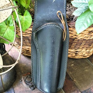 May include: A black leather holster with a stitched design, featuring a loop and a leather cord. The holster is set against a backdrop of greenery and a vintage wooden trunk, suggesting a Western or historical theme.