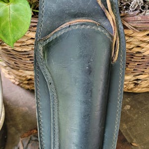May include: Black leather holster with stitched detailing and a leather tie. Designed to hold a handgun. The holster is set against a backdrop of foliage and a wooden surface.