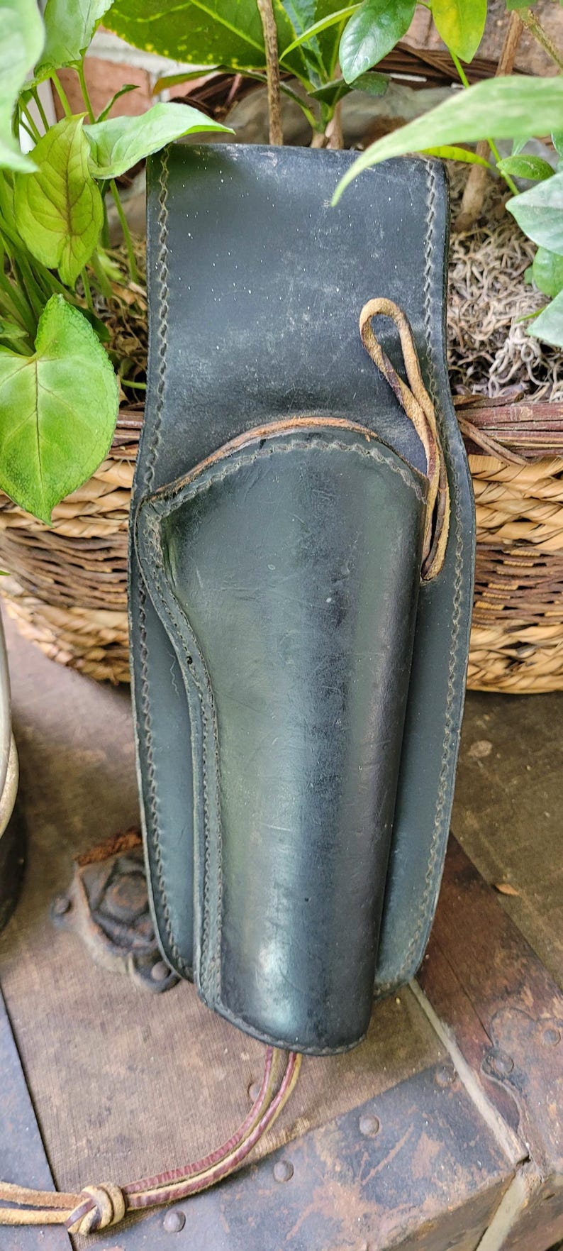 May include: Black leather holster with stitched detailing and a leather tie. Designed to hold a handgun. The holster is set against a backdrop of foliage and a wooden surface.
