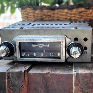 May include: Classic Chevrolet car radio with chrome knobs and a black display showing radio frequencies. The radio is silver and rectangular. It rests on a wooden trunk. The radio's face has the Chevrolet logo.