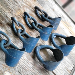 May include: Six blue metal objects with a chain-like design, on a weathered wooden surface. Each object has a loop and a small, cup-like structure. The objects are arranged diagonally across the frame.