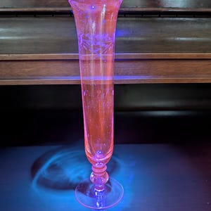 May include: A tall, slender, pink glass bud vase with a flared rim and a circular base. The glass features an etched floral pattern around the upper section and exhibits a bright, glowing pink fluorescence under ultraviolet light.