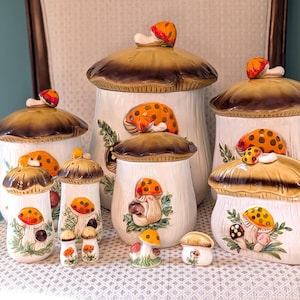 May include: A collection of ceramic mushroom-shaped kitchen canisters and accessories. The canisters have brown and tan lids with orange and yellow mushroom accents. The white canisters are decorated with painted mushroom and leaf designs.