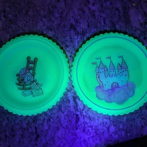 May include: Two round, glowing green plates with scalloped edges. One plate features a rocking chair with cartoon animals and books. The other plate has a castle design with flags. The plates are illuminated with a black light.