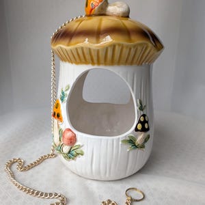 May include: A white ceramic mushroom-shaped bird feeder with a brown and tan cap, featuring painted mushroom and leaf accents. It has a gold chain for hanging and a small ring at the end. The feeder has a large opening for birds to enter.