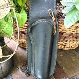 May include: Black leather holster with stitched detailing and a leather tie. The holster is placed on a vintage wooden trunk, with plants in the background. The holster is designed to hold a handgun.